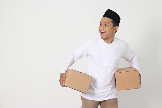 Portrait of happy Asian muslim man in koko shirt with peci looking confident while carrying cardboard box. Homecoming on Eid Mubarak concept. Isolated image on white background