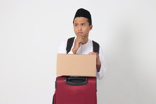 Portrait of confused Asian muslim man holding suitcase and cardboard box and thinking with hand on chin. Going home for Eid Mubarak. Isolated image on white background