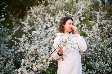 Woman allergic suffering from seasonal allergy at spring in blossoming garden at springtime. Young woman sneezing, closing nose by hand in front of blooming tree. Spring allergy concept