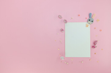 Easter party concept. Flat lay, top view of blank sheet of paper, easter bunny, multicolored candy eggs, small flowers on isolated pastel pink background