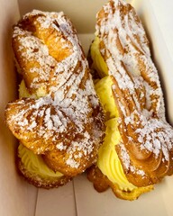 close in a cardboard box lie two eclairs with custard sprinkled with powdered sugar. sweets. food with a lot of calories