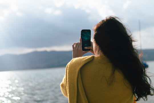 A young woman in a yellow scarf takes a photo of a lake on her phone, with the sun setting behind the mountains