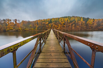 Beautiful view to long steel and wood bridge over big and silent lake with autumn golden trees and bushes around at cloudy and rainy morning