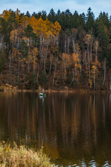 Beautiful view to the coast of big lake with beautiful high green and golden trees at fall reflecting in water with fisherman on small boat at the center