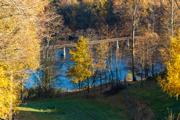 Beautiful mountain landscape full of golden and green trees at fall and long steel and wood bridge at the center of big lake at down of the hill