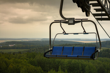 empty four-seater open cabin of cable car chairlift with blue seats moves over hilly forest against cloudy sky