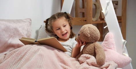 Cute little girl with toy bear reading story in bedroom at night