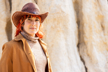 A woman wearing a brown hat and a brown coat stands in front of a rock wall. She has ared hair and...