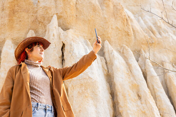 A woman in a brown coat and hat is taking a selfie in front of a rocky cliff. The photo has a...