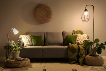 Stylish grey sofa with pillows, ottomans, lamps and houseplants near beige wall at night
