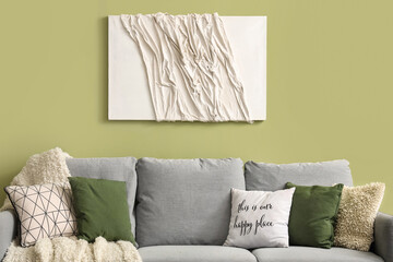 Stylish grey sofa with pillows, blanket and painting near green wall