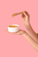 Female hands holding container with sugaring paste and spatula on pink background