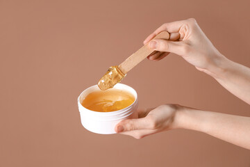 Female hands holding container with sugaring paste and spatula on brown background