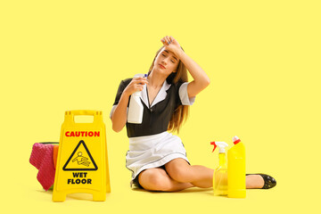 Portrait of tired female janitor with bottles of detergent and caution sign on yellow background
