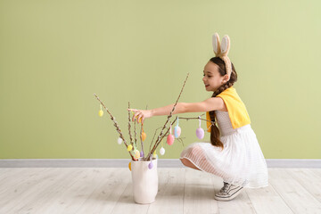 Cute little girl in bunny ears decorating willow branches with Easter eggs on green background - obrazy, fototapety, plakaty