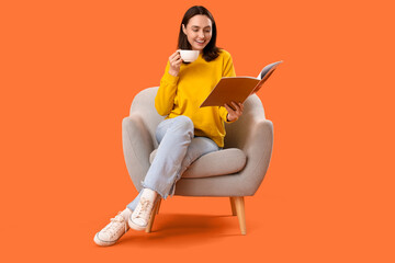 Young woman with cup of tea reading magazine in armchair on orange background