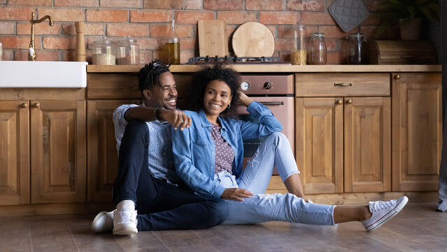 Attractive African couple sit on floor in kitchen smiling feel satisfaction resting at relocation day move at own house, happy clients of remodelling and repair services, dating, bank mortgage concept
