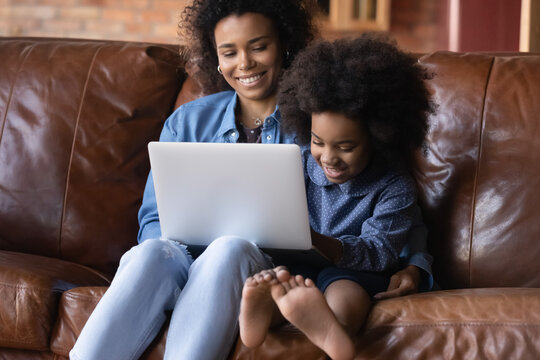 African young mother and little daughter sit on sofa with wireless computer, caring mom teach kid using laptop for education or fun, family choose goods buying toys on internet, modern tech concept