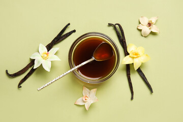 Vanilla extract in bowl with spoon, vanilla pods and flowers on green background. Top view