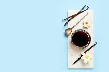 Vanilla extract in bowl with spoon, vanilla pods and flowers on blue background. Top view