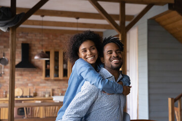 Happy beautiful African wife piggyback cuddling husband, family smiling looking at camera, posing inside modern cottage house. Vacation, celebrate relocation day, romantic relations, tenancy concept