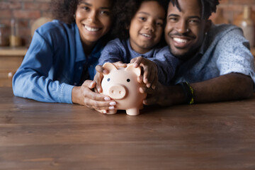 Happy African couple little daughter sit at table with piggybank smile look at camera, close up focus on money box. Deposit, make financial investment, save money for future, for kid education concept