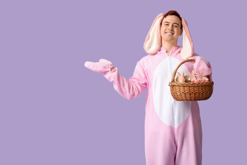 Happy young man in Easter bunny costume holding basket and pointing at something on lilac background