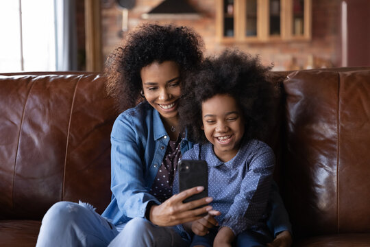 Happy African 6s girl and young mother sit on sofa with smartphone, enjoy video conference talk event with family, smiling watching funny online content, amusing videos. Modern technology, fun concept