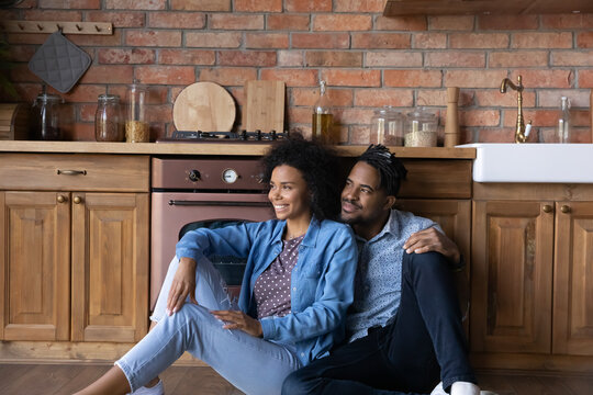 African ethnicity couple sit on floor in kitchen having dreamy mood, smile look into distance, daydream together about bright future in new own home. Bank loan, remodelling, repair services concept
