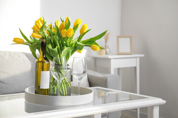 Beautiful vase of yellow tulips with bottle of wine and glasses on coffee table in living room