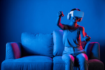 Smart Female sitting on sofa surrounded by neon light wear VR headset connecting metaverse,...