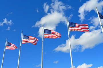 multiple American flags flying on poles against a bright blue sky with fluffy white clouds.