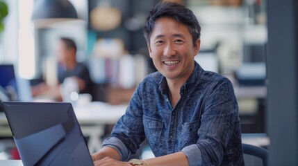 an Asian man working on a laptop at the office, smiling, looking at the camera