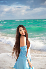 Fototapeta na wymiar A beautiful brunette with long hair in a blue dress poses against the background of the ocean. Vacation