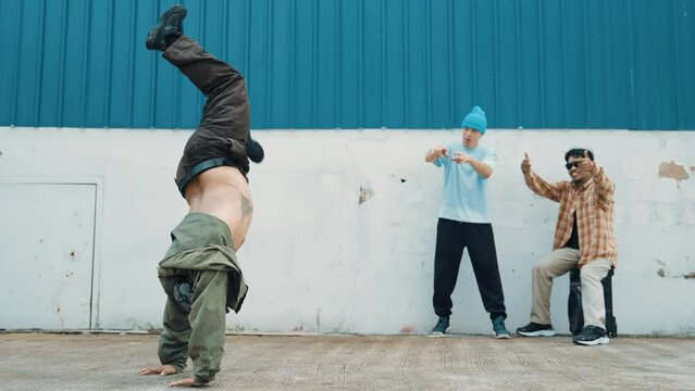 Skilled hipster doing cart wheel while multicultural friends encourage him. Hip-hop dancer moving foot step to music surrounded by group of hipster at wall near street. Outdoor sport 2024. hiphop.