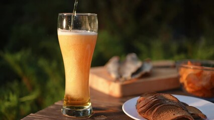 Close-up. Slow motion. Beer added to a beer glass and foam flows down the glass. A beer glass with beer, crackers, chips in a glass bowl and salted dried fish on a wooden board stands on a wooden