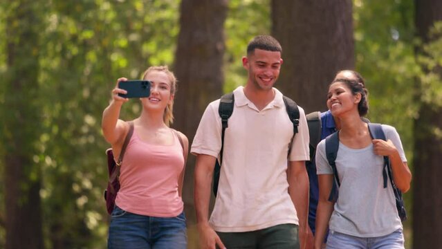 Young couple with friends wearing backpacks hiking along trail posing for selfie on mobile phone whilst walking in summer forest countryside - shot in slow motion