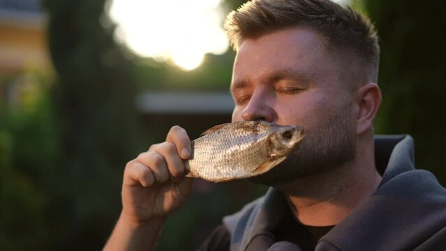 Slow motion. Close-up. A young bearded man sniffs salted dried fish that he holds in his hand. The man closes his eyes and enjoys the smell while standing outdoors