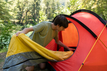 Happy teenager preparing camp and storing his sleeping bag in the tent before going out to enjoy...