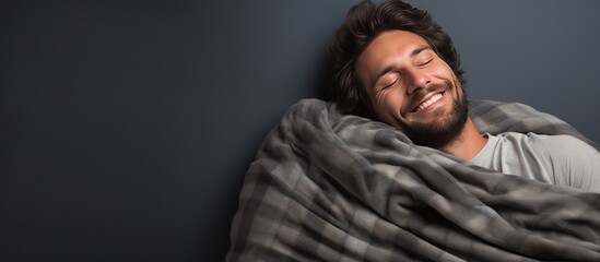 A handsome man is smiling while wrapped up in a soft blanket and holding a pillow on a grey background.