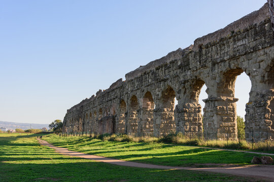 The long arched wall of an ancient Roman aqueduct along a walking path in a park, evening shadows from the setting sun shadows fall on the ground in a beautiful pattern