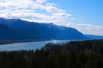 View down a massive river with high mountains in the background and pine trees in the foreground on a partially clear day. 