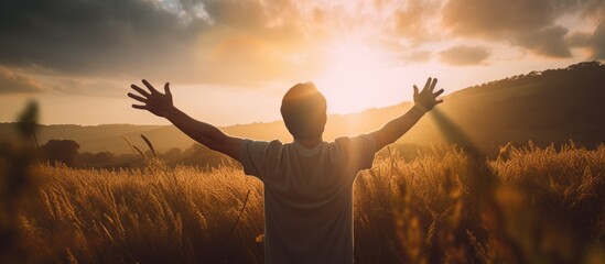A young man is standing in a field during the evening, reaching his arms out towards the sunlight.
