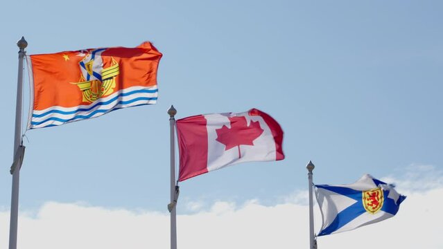 Close up of the Acadian flag, the Canadian flag and the Nova Scotia flag in a cascading row and blowing in the wind, against a blue sky with clouds, on a windy summer day