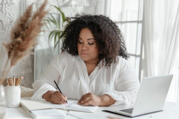 Plus-size young woman concentrating on writing, with a laptop on her desk. - obrazy, fototapety, plakaty