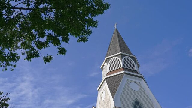Low angle of old wooden church steeple with a cross and tree branches in the foreground, against a blue sky, on a breezy summer day