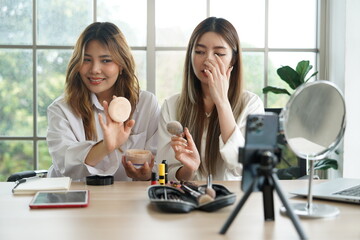 Two Asian girls, partners and roommates sell cosmetics online, introduce powder, lipstick, brushes, use smartphones and laptops to work in an apartment.
