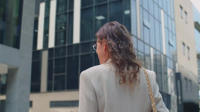 Back tracking shot of cheerful female entrepreneur turning back to camera while walking at business district