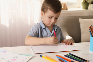 Cute little boy drawing with pencil at white wooden table in room. Child`s art