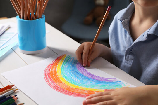 Little boy drawing rainbow with pencil at white wooden table indoors, closeup. Child`s art